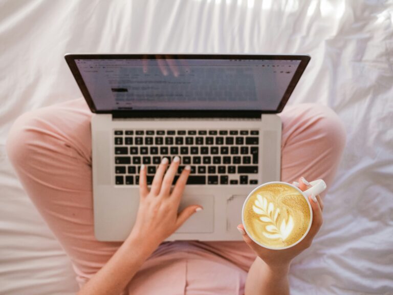 woman seated cross legged with laptop across knees and cup of coffee in one hand
